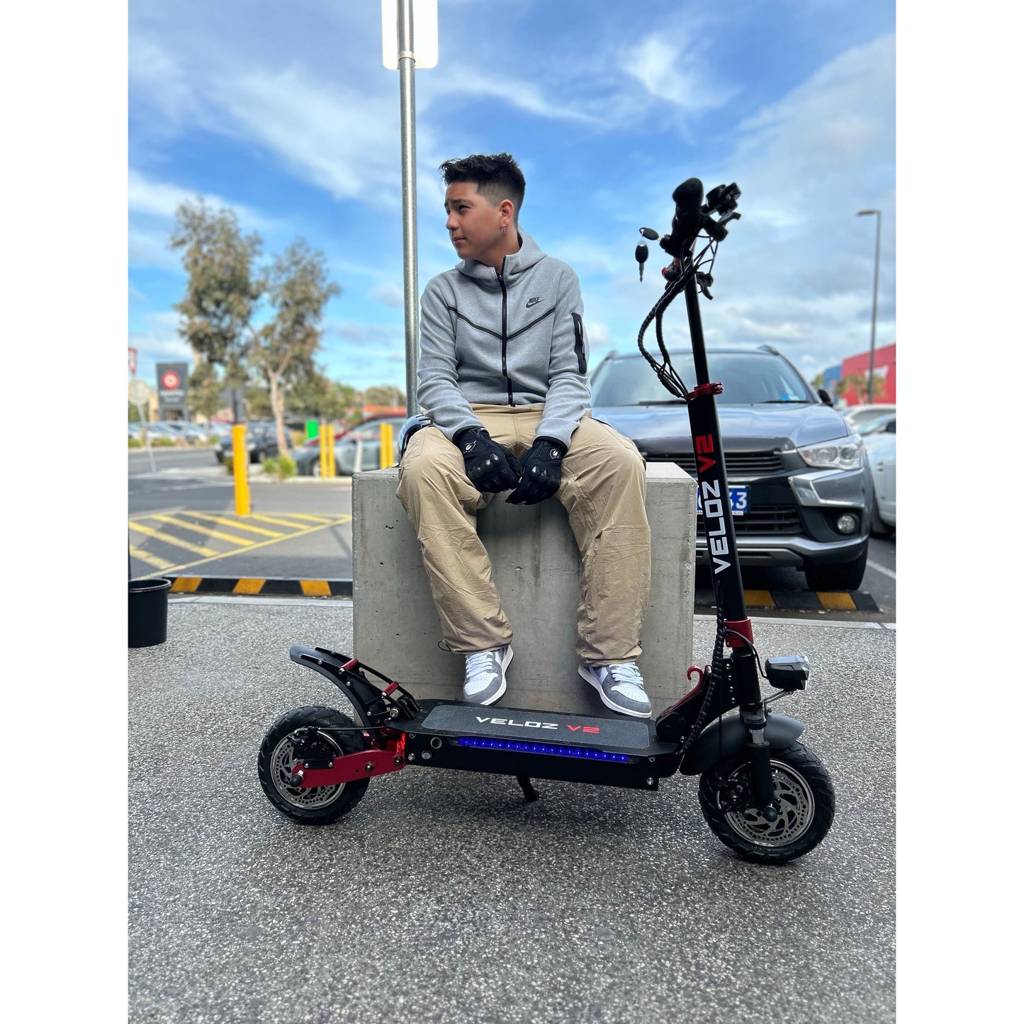 a teenage boy sitting next to his parked veloz v2 electric scooter in parking lot